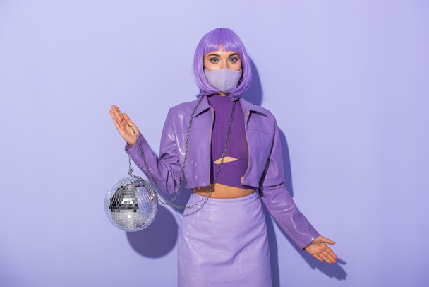 young woman dressed in doll style in medical mask with disco ball on violet colorful background - Photo, Image