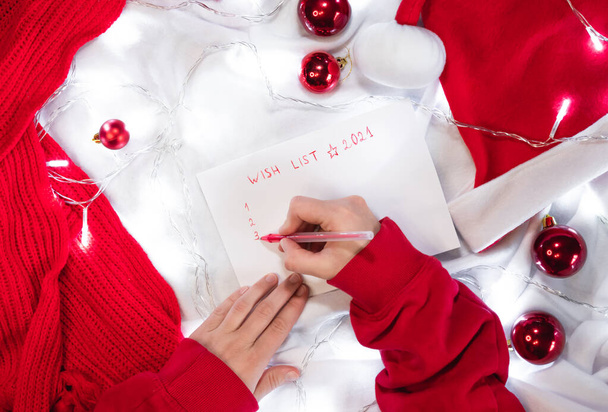 Escribe una lista de deseos para Santa con un bolígrafo rojo en una hoja blanca en blanco. Bolas rojas de Navidad, sombrero de Santa y bufanda hecha a mano. Escribe una carta con deseos en una manta blanca. - Foto, imagen