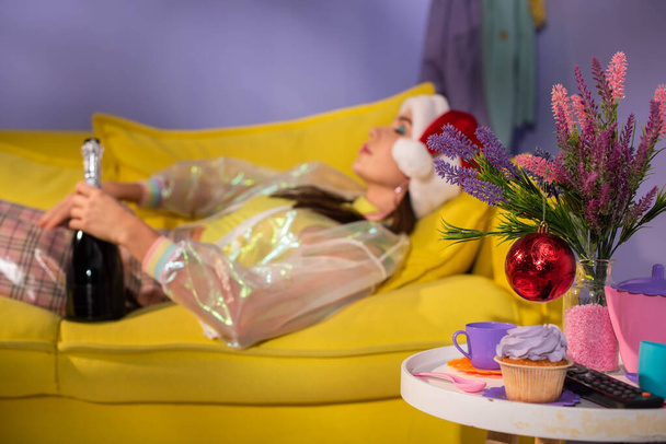 Різдвяні прикраси та молода жінка в капелюсі Санти позує як лялька на жовтому дивані з шампанським
 - Фото, зображення