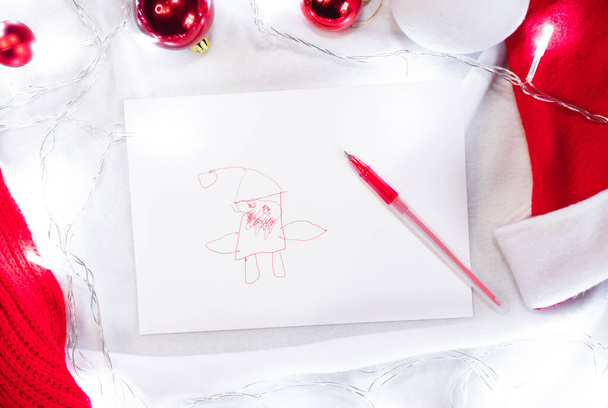 Kreatywny dziecięcy rysunek Świętego Mikołaja na białym tle. Cute Santa narysowane przez dziecko z czerwonym piórem i białym arkuszem papieru i wokół są czerwone kulki świąteczne i światła girlanda. - Zdjęcie, obraz