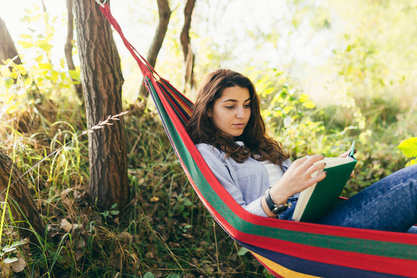 Giovane bella ragazza che riposa in un'amaca colorata nel parco e legge un libro - Foto, immagini