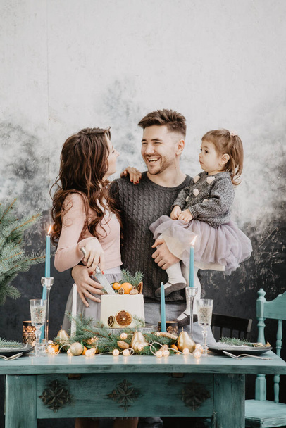 Young family with a daughter in festive outfits at a served table with candles, garlands, sparklers and a cake near the Christmas tree - Foto, Bild