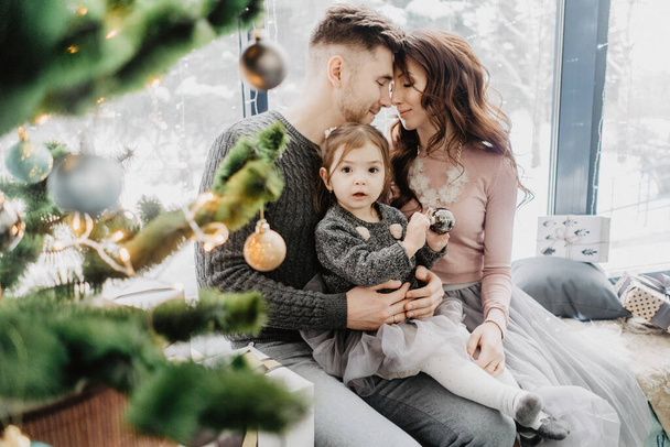 Young family with a daughter in festive outfits with a garland near the Christmas tree - Foto, Bild