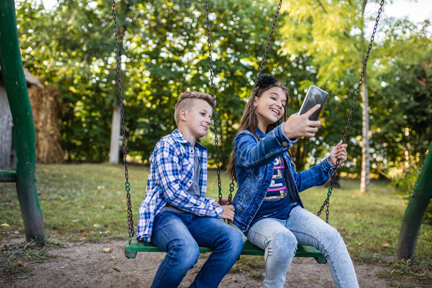 Little boy and girl are taking picture of themselves for selfie with phone while sitting on swing - Photo, image