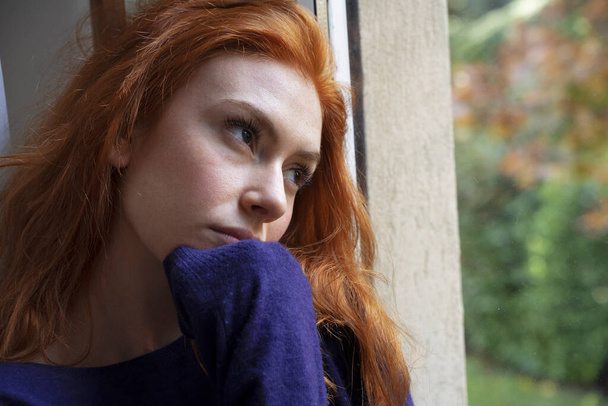 Νεαρή ανήσυχη γυναίκα κοιτάζει έξω από το παράθυρο - Φωτογραφία, εικόνα