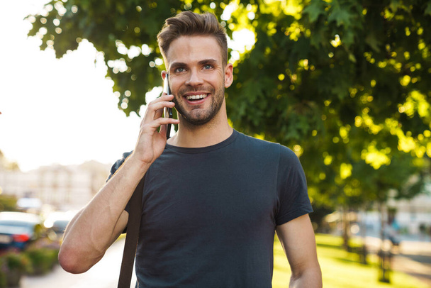 Smiling attractive young man talking on mobile phone while walking on a street - Foto, Imagem