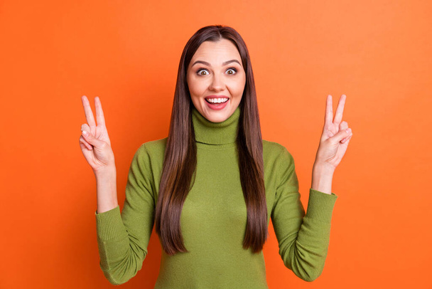 Photo of young attractive smiling excited happy girl showing v-sign wear green turtleneck isolated on orange color background - Foto, afbeelding