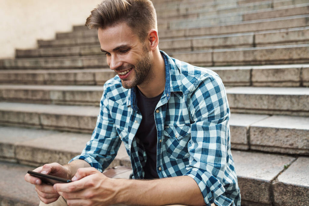 Handsome smiling young man using mobile phone while sitting on steps outdoors - Fotó, kép