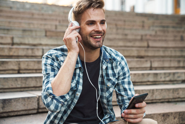 Attractive man in headphones holding mobile phone while sitting on steps outdoors - 写真・画像