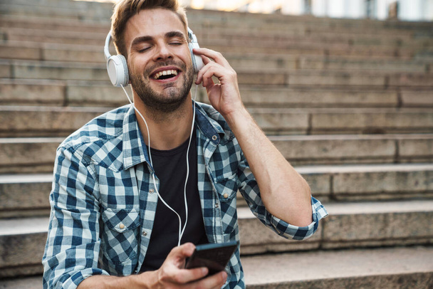 Attractive man in headphones holding mobile phone while sitting on steps outdoors - Foto, Imagem