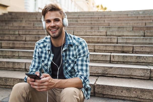 Handsome man sitting on stairs with headphones and mobile phone outdoors - Fotó, kép