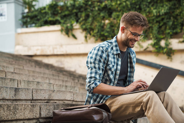 Young smiling man sitting on steps and working on laptop outdoor. Freelancer - Foto, Imagen