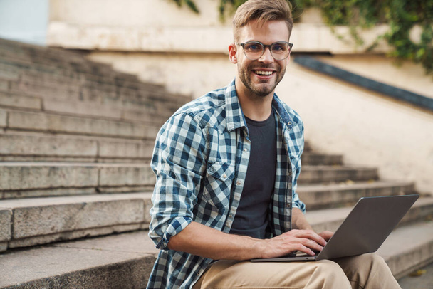Fiatal férfi egy laptop az utcán, ül a lépcsőn dolgozik az interneten, és kommunikál a barátaival videóhívás - Fotó, kép