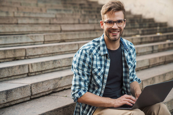 Young smiling man sitting on steps and working on laptop outdoor. Freelancer - Photo, image
