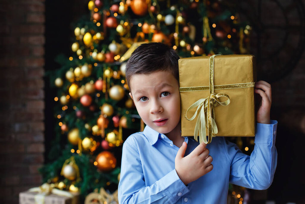 Söpö pieni poika noin viisivuotias lahja koristeltu joulukuusi. Yllättynyt, hän laittoi lahjan korvaansa. - Valokuva, kuva