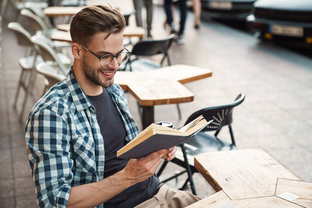 Smiling man enjoying free time and reading a book while sitting outdoors at coffee shop - Foto, Imagen