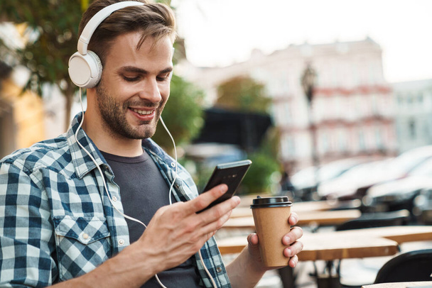 Smiling young man wearing headphones sitting at the cafe table outdoors, using mobile phone - 写真・画像