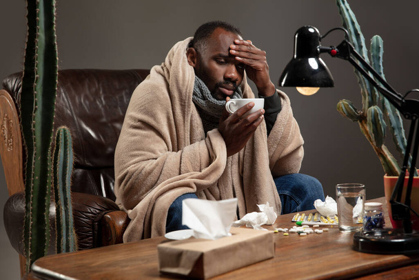 Young African-american man wrapped in a plaid looks sick, ill, has terrible headache sitting at home indoors. - Foto, imagen