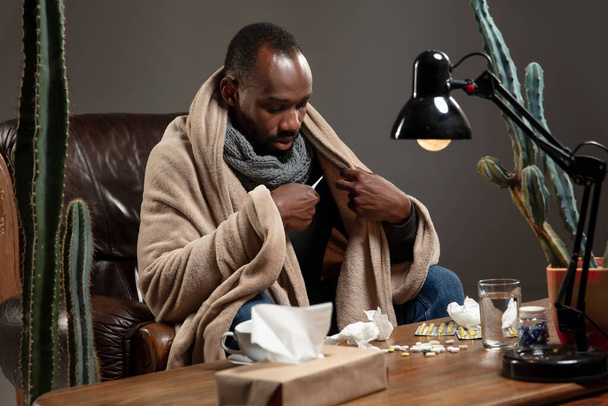 Young African-american man wrapped in a plaid looks sick, ill, sitting at home indoors. Healthcare concept - Foto, Imagen