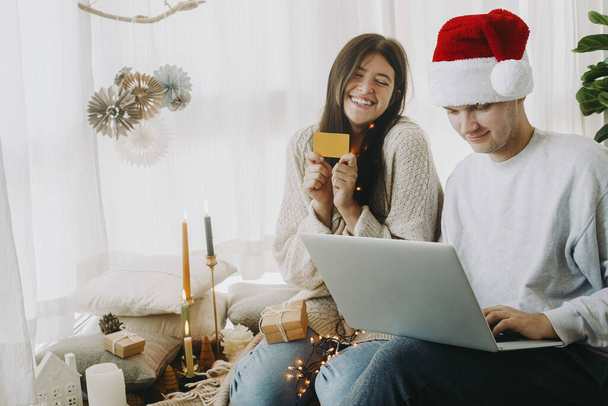 クリスマスセールやオンラインホームショッピング。ブラックフライデーにラップトップとクレジットカードのオンラインショッピングを使用して幸せなスタイリッシュな家族は、お祝いの休日の装飾が施されたモダンな部屋に座っています。興奮した妻 - 写真・画像