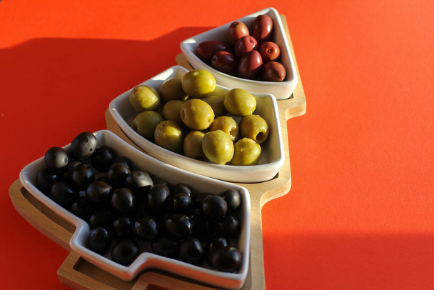 Green, black and red olives in a bowl in the shape of a Christmas tree on a red background. Concept of new year's dishes, Christmas celebrations and Mediterranean dishes. Top view, copy location. - Photo, Image