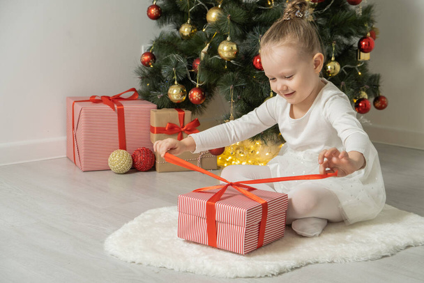 Cheerful cute child girls opening gift box having fun near christmas tree wearing white dress. - 写真・画像
