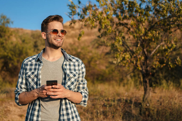 Handsome happy guy smiling and using cellphone while strolling on nature at summer - Foto, Bild