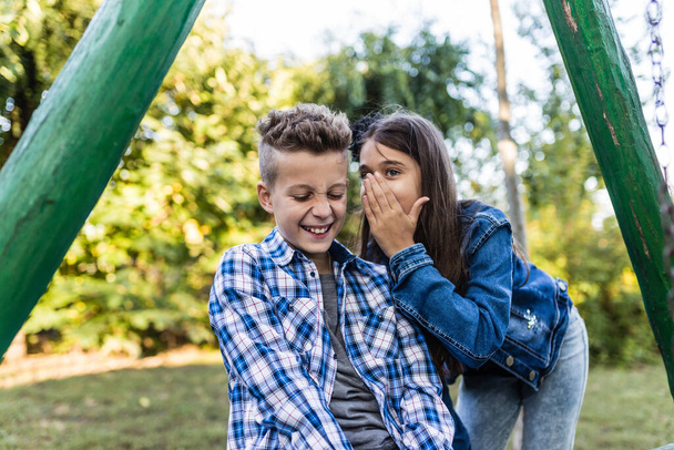 Zavřít dva šťastní malí přátelé se smějí a baví v parku. Pozitivní děti si šeptají tajemství do uší.. - Fotografie, Obrázek