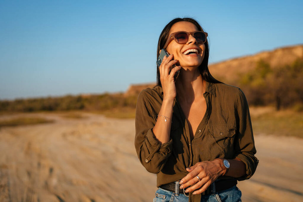 Beautiful laughing girl talking on cellphone while strolling on nature at summer - 写真・画像