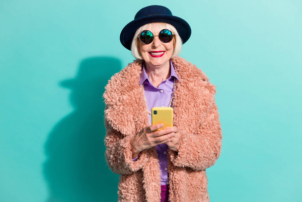 Photo of cheerful senior lady wear retro jacket headwear eyeglasses holding device isolated teal color background - Zdjęcie, obraz
