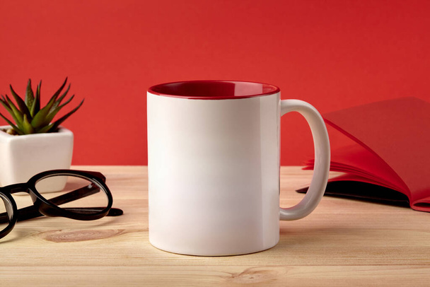 White porcelain mug on wooden desktop next to notepad, glasses and green succulent in pot against red studio background. Close up, copy space - Φωτογραφία, εικόνα
