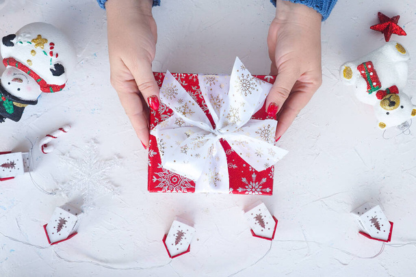Frauenhände halten eine rote Schachtel mit einem Weihnachtsgeschenk, einem Kranz - Häuser und ein niedlicher Weihnachtsbaum spielen einen Schneemann und einen Bären auf einem weißen Tisch. Blick von oben. Lebensstil - Foto, Bild