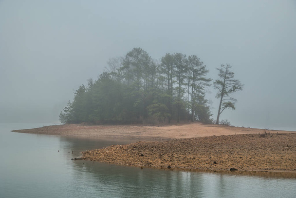 Thick morning fog at the lake with the woodlands on a small island obscured by the conditions of the weather in wintertime - Photo, Image