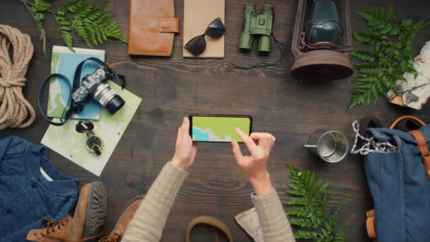 Top view of unrecognizable woman holding mobile phone over wooden table with various travel accessories lying on it and using digital map - Footage, Video