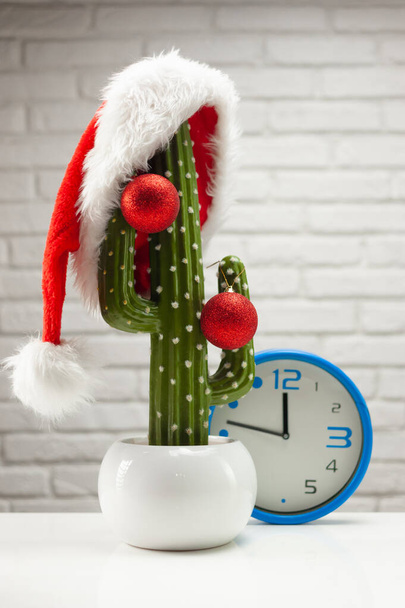 サンタクロースの帽子と光の背景にクリスマスのおもちゃを持つ人工サボテン - 写真・画像