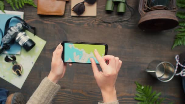 Top view zoom out of unrecognizable woman using map on mobile phone. Photo camera, rope, backpack, binoculars, lantern and other travel gear lying on wooden table decorated with fern and birch logs - Footage, Video