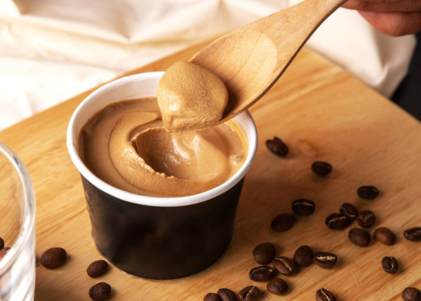 Crop image of hand scooping coffee ice cream in a paper cup with a wooden spoon on a wooden cutting board with messy coffee beans decoration. - Foto, Imagen