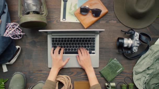 Top view zoom out of unrecognizable woman planning trip on laptop standing on wooden table with various travel accessories lying on it - Footage, Video