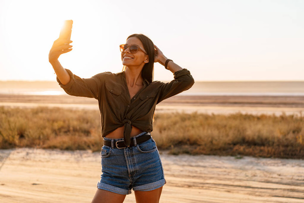 Beautiful joyful girl taking selfie on cellphone while strolling on nature at summer - Фото, зображення