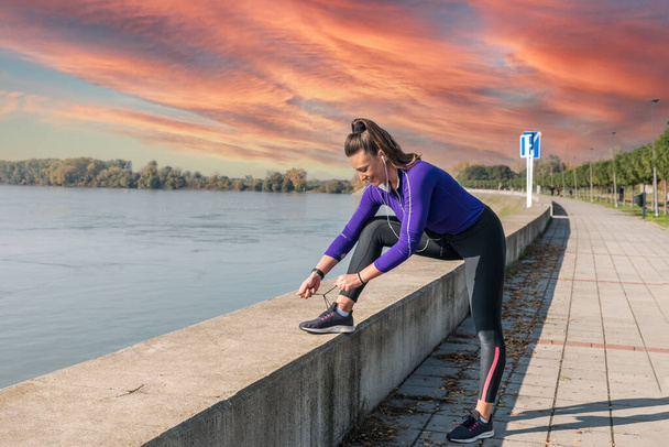 Young active sporty runner woman ties a shoelace before she start her running exercise to jogging by the river with beautiful sunset - Φωτογραφία, εικόνα
