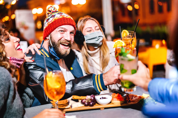 Erkekler ve kızlar gece kokteyl barında yüz maskesi takarak süslü içecekler içiyorlar - yeni normal yaşam tarzı konsepti, dışarıda sarhoş arkadaşların birlikte eğlenmesi - canlı filtre, kış şapkalı adama odaklandı - Fotoğraf, Görsel