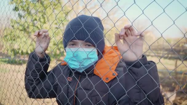 Portret van schattige ongelukkige kleine jongen in medisch masker op kinderen speeltuin achter metalen gaas in zelfisolatie - Video