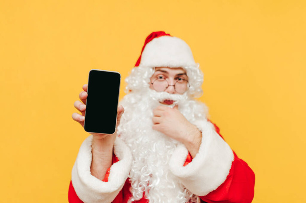 Le Père Noël attire l'attention en montrant un smartphone, tenant un appareil dans sa main, regardant intéressé. Focus sur un téléphone portable. - Photo, image