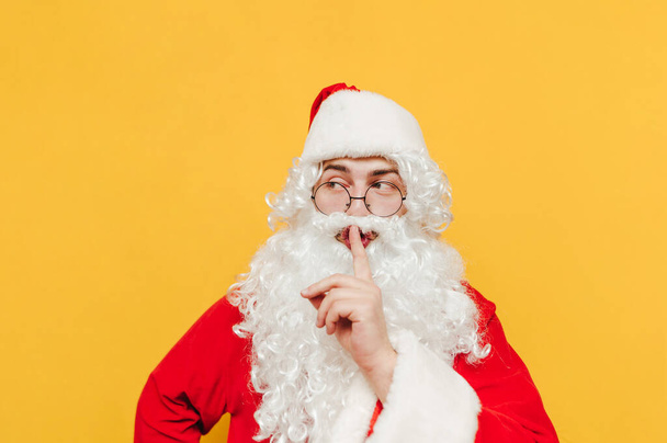 サンタクロースの衣装の男は、秘密を隠して、静かなジェスチャーを表現しています。サンタは彼の唇に指で触れることによって急いでジェスチャーを示しています. - 写真・画像