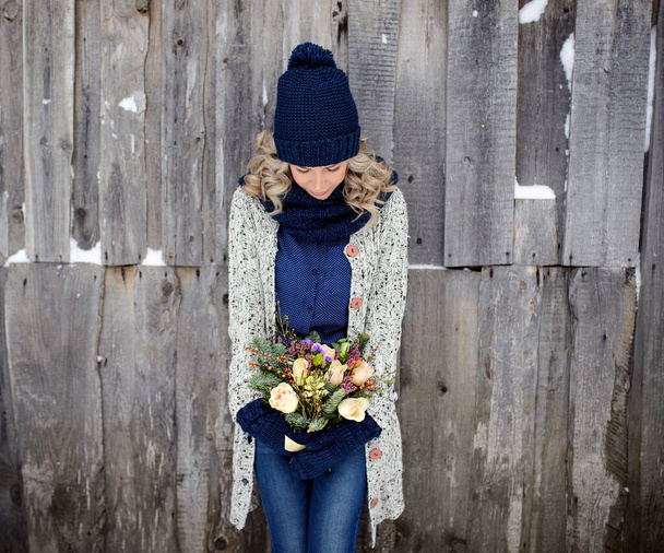 An attractive woman holds a bouquet of roses and fir branches against a wooden wall. Stylish winter clothing - Φωτογραφία, εικόνα