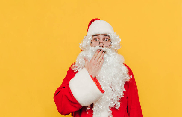 Překvapený Santa Claus se dotýká úst rukou ukazující udivené gesto, je v šoku, dívá se na kameru s vystrašenýma očima dokořán otevřenýma.. - Fotografie, Obrázek