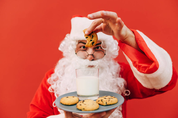 Vtipná fotka veselého starého Santa Clause, jak drží talíř se sušenkami na ruce, bere jednu sušenku a dává ji do sklenice mléka. Zaměření na cookie, rozmazané pozadí. - Fotografie, Obrázek