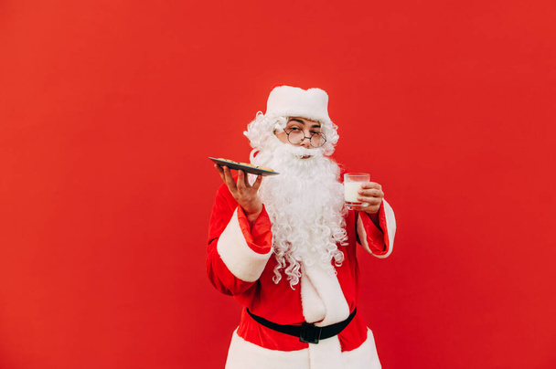 Санта-Клаус в модном костюме и очках позирует для фото на красном фоне и держит тарелку с печеньем и стаканом молока. - Фото, изображение