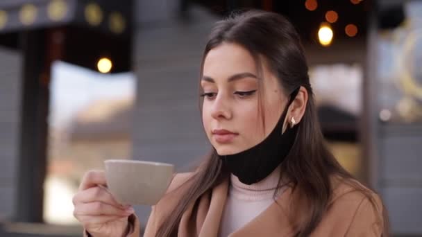 Процесс красивой женщины снимает защитную маску для питья кофе. Женщина сидит на террасе и пьет кофе. Карантин, самоотдаление. Ковид-19 - Кадры, видео