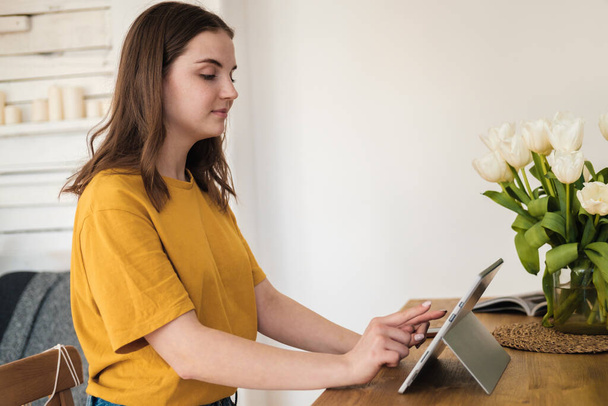 Young beautiful woman in yellow t-shirt usimg tablet for work, online business, studying sitting at home. Self-isolation, study, work during quarantine, pandemic. Remote job, education, student. - Φωτογραφία, εικόνα